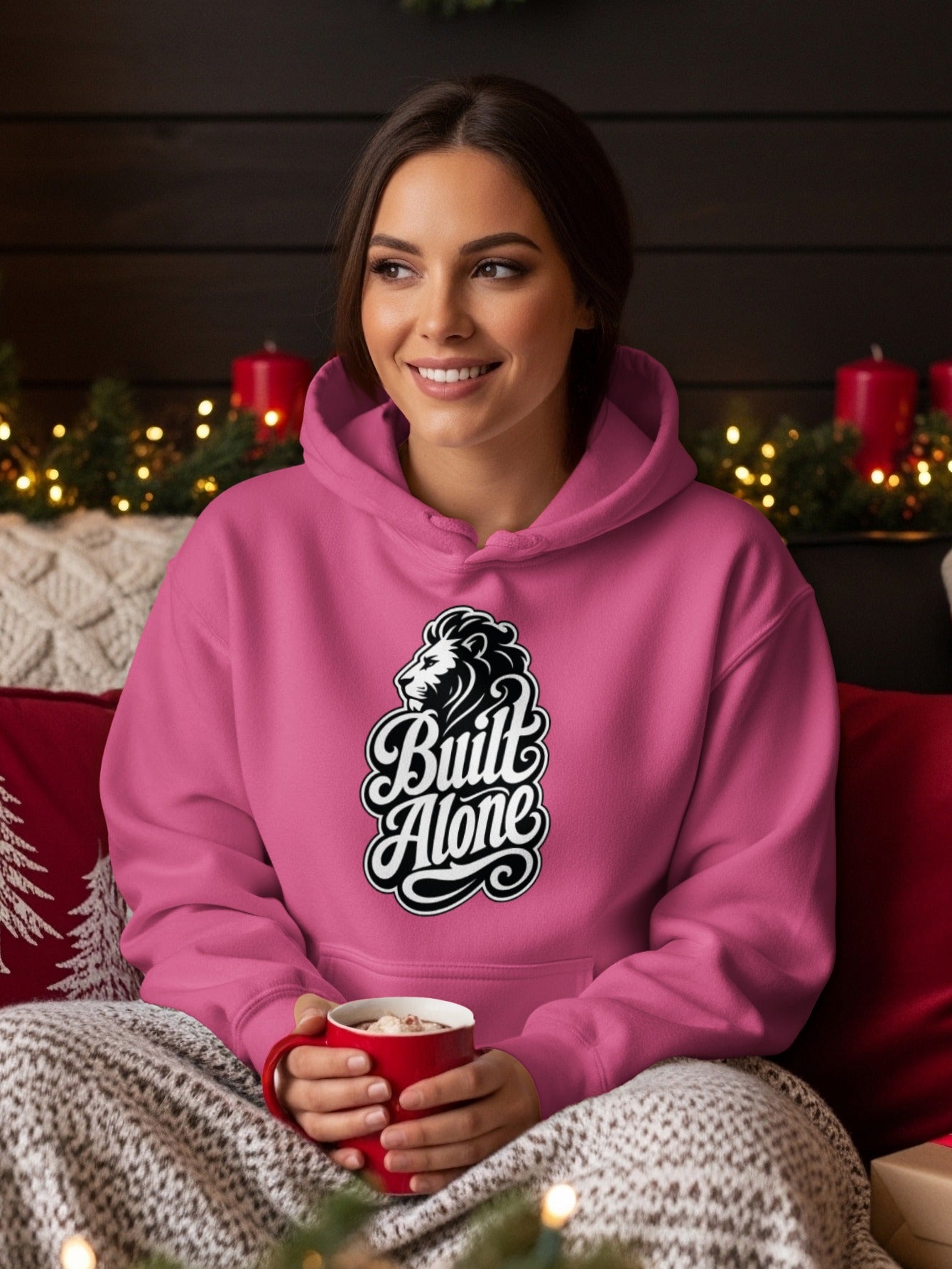 Pink hoodie with "Built Alone" text and lion logo, held by a woman with a mug.
